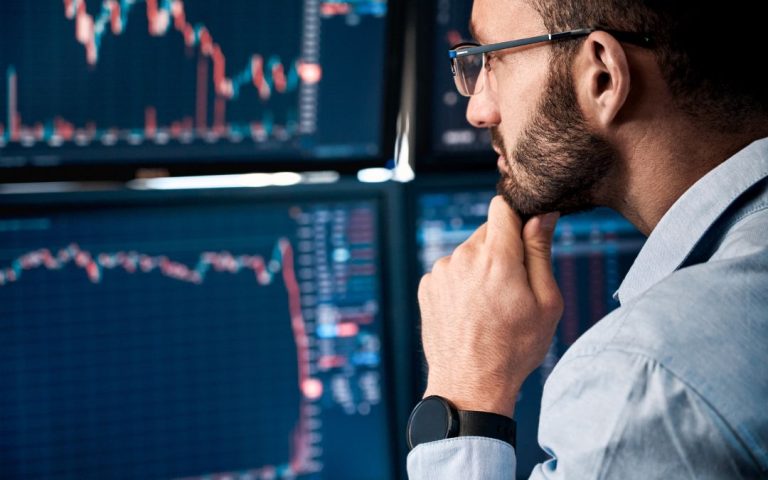 bearded man in glasses thoughtfully staring at trading charts while holding his chin